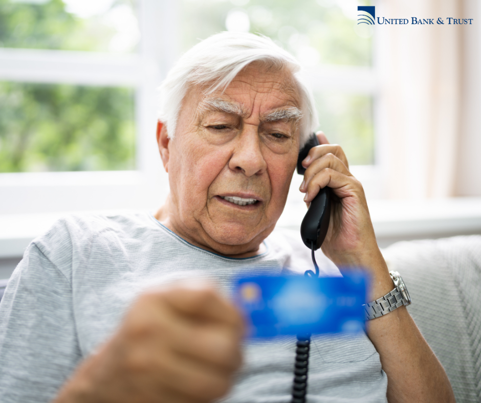 Older man speaking into a corded phone while squinting at the debit card he holds in front of him.